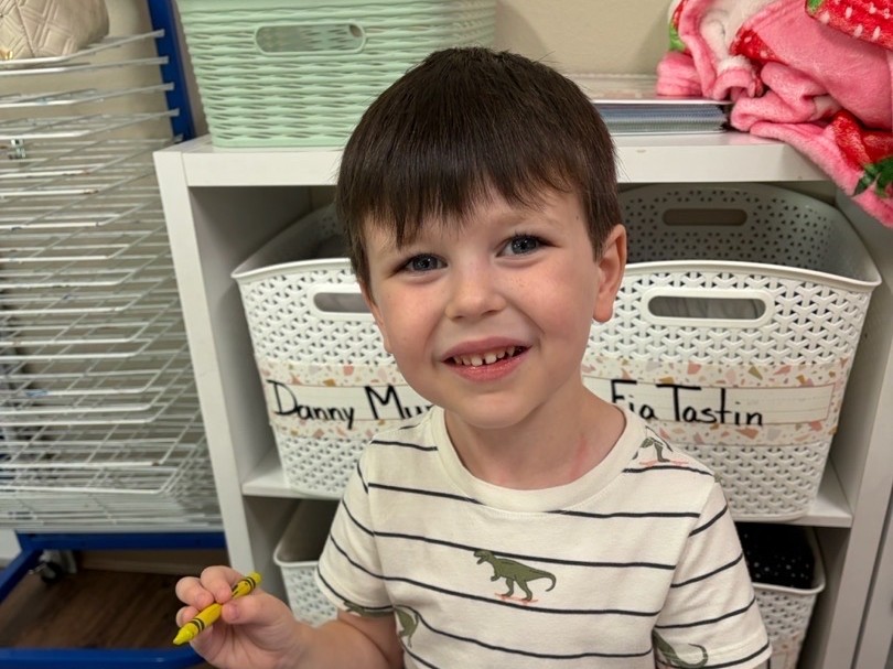 A smiling preschool boy in a dinosaur shirt holds up a yellow crayon beside his workbook at the classroom table.