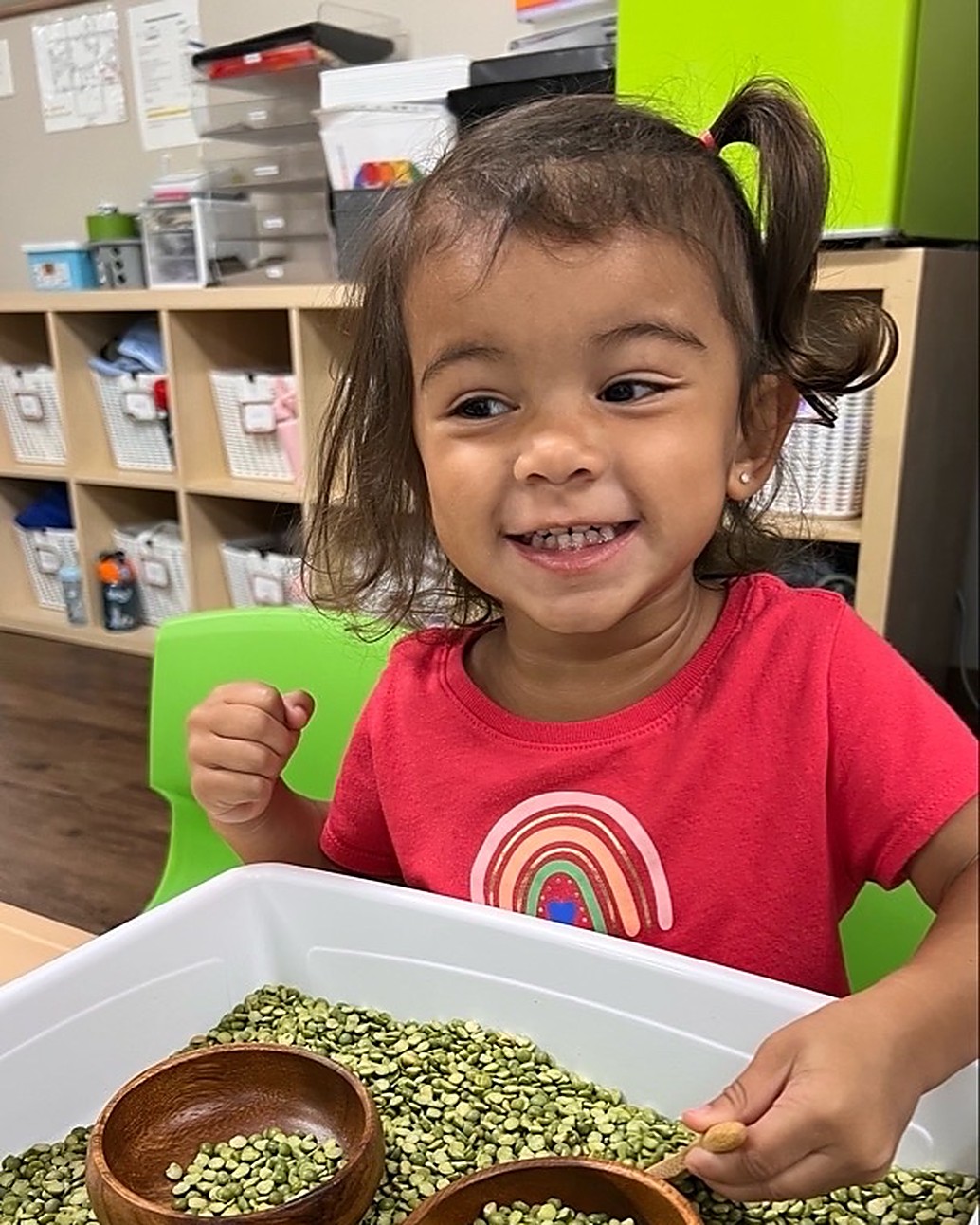 A smiling toddler with pigtails in a red shirt scoops green split peas with a wooden spoon at a classroom sensory table.
