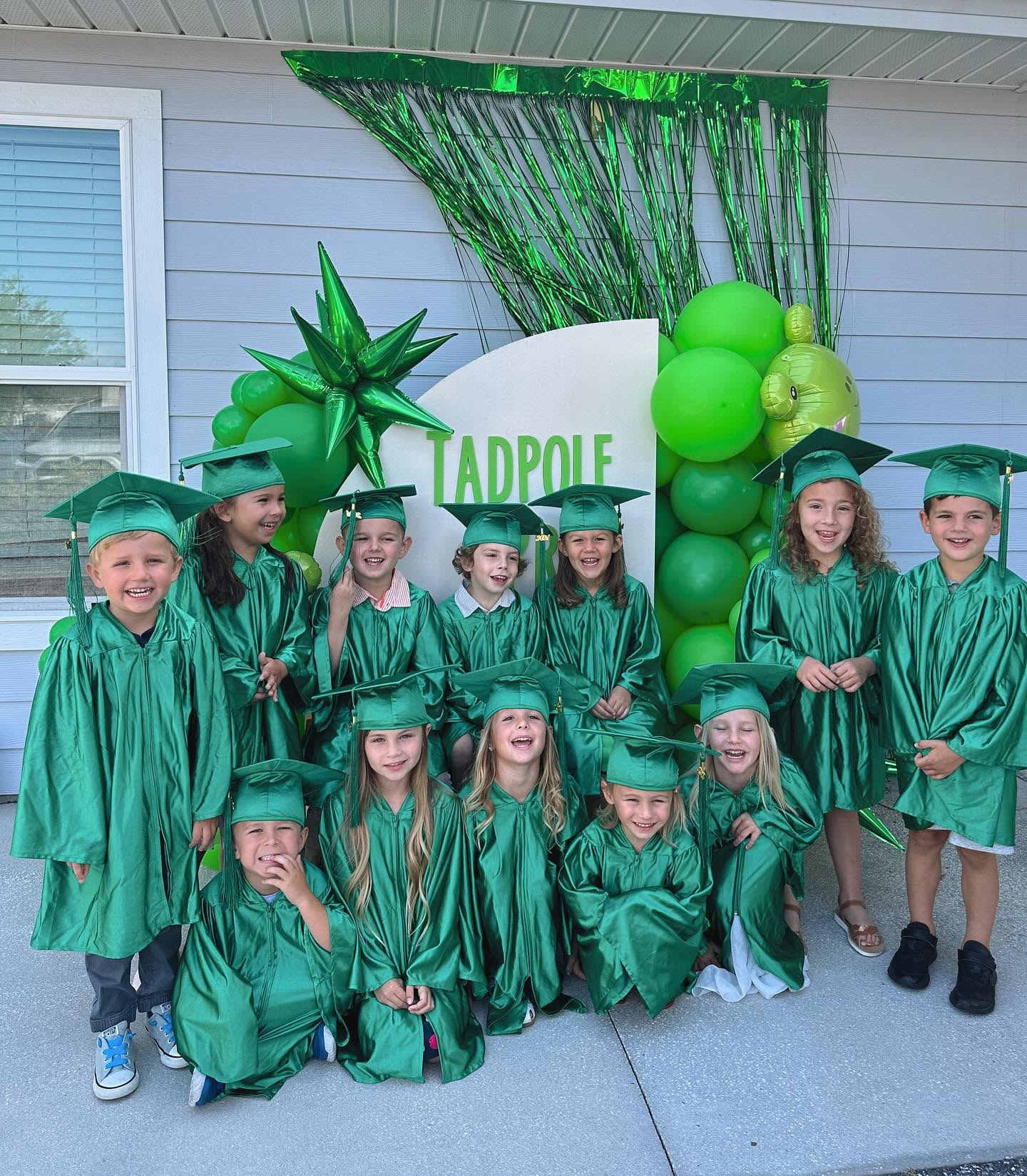 Preschool graduates in green caps and gowns gather smiling in front of a Tadpole Prep celebration backdrop.