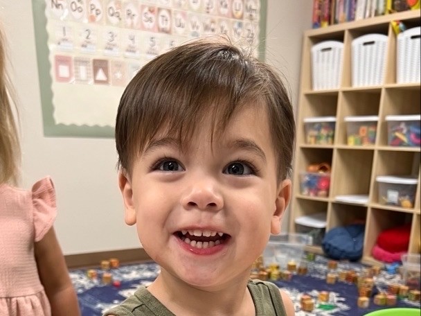 A happy toddler in a green tank top shows his red-paint-covered hand beside his handprint painting.