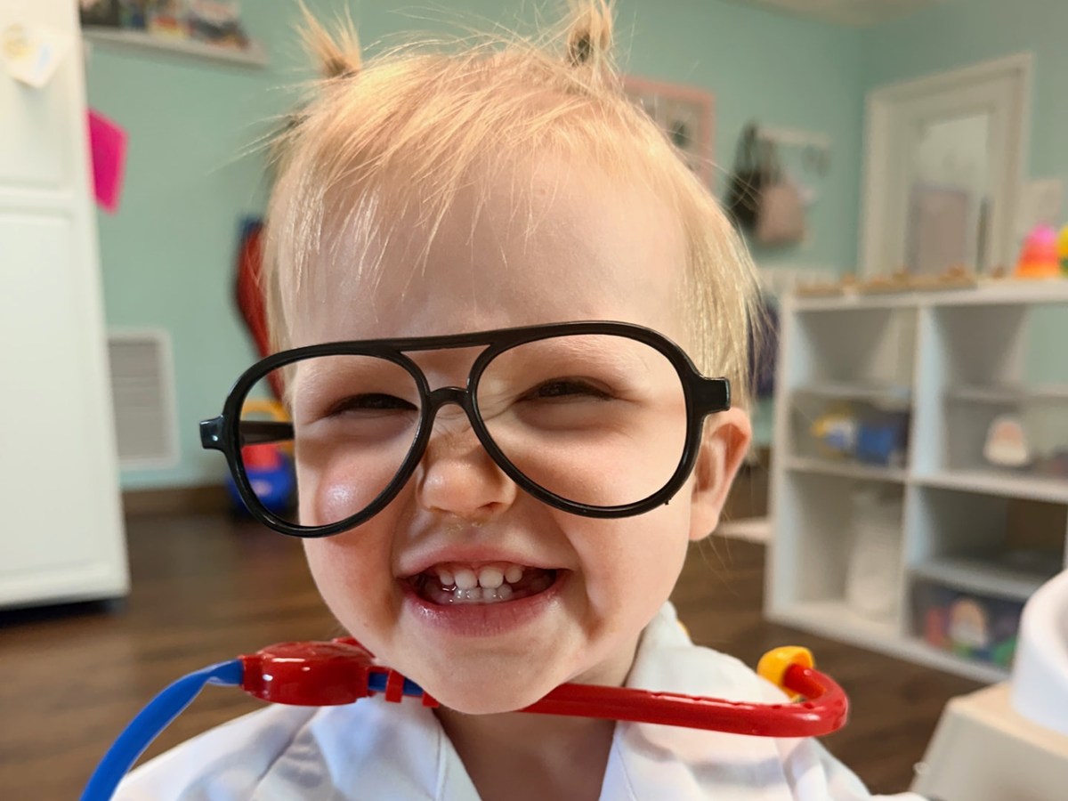 A smiling toddler dressed as a doctor, wearing oversized glasses and a toy stethoscope.